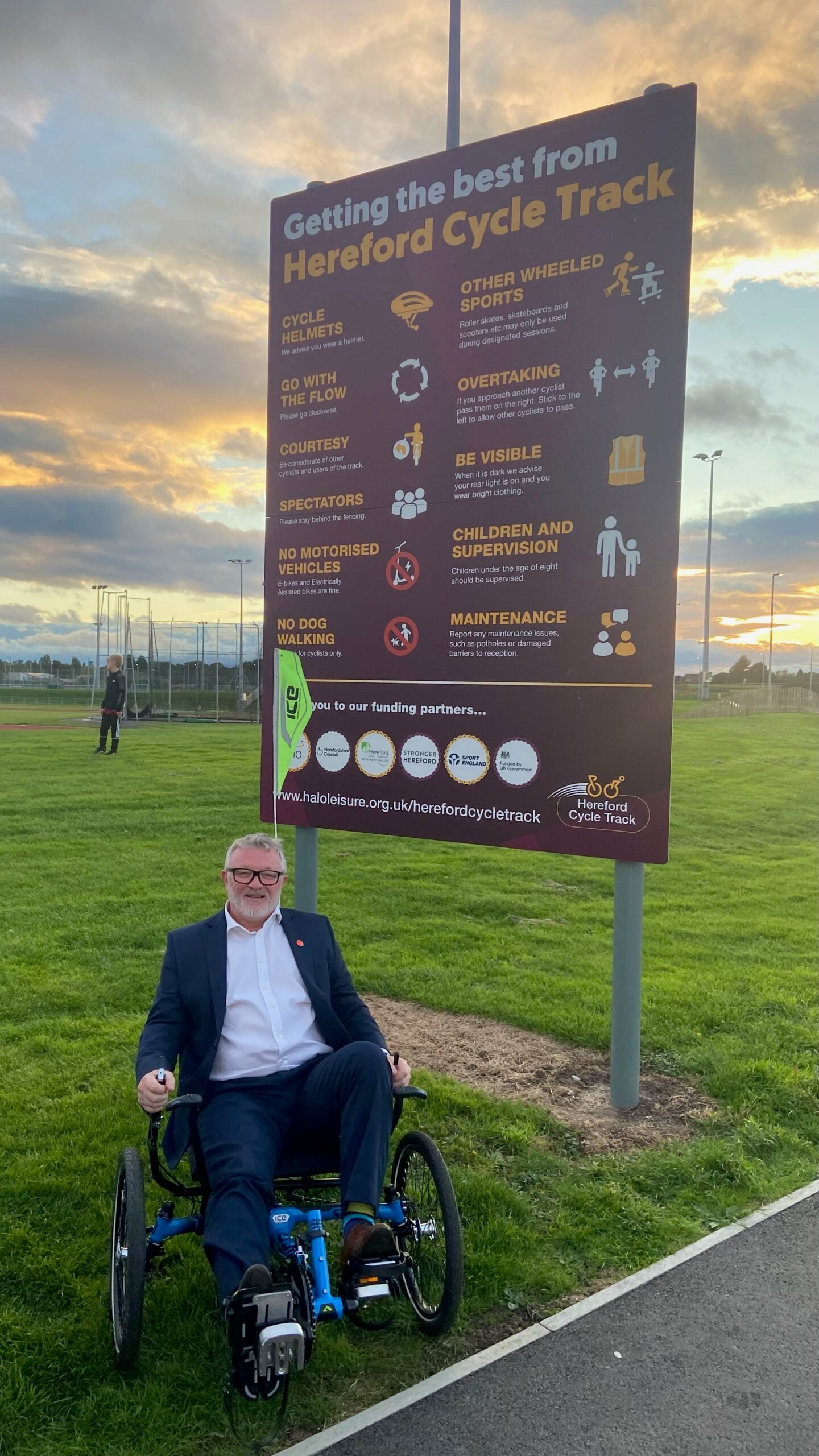 Large outdoor information sign about Hereford Cycle Track with a man seated in a trike by the sign