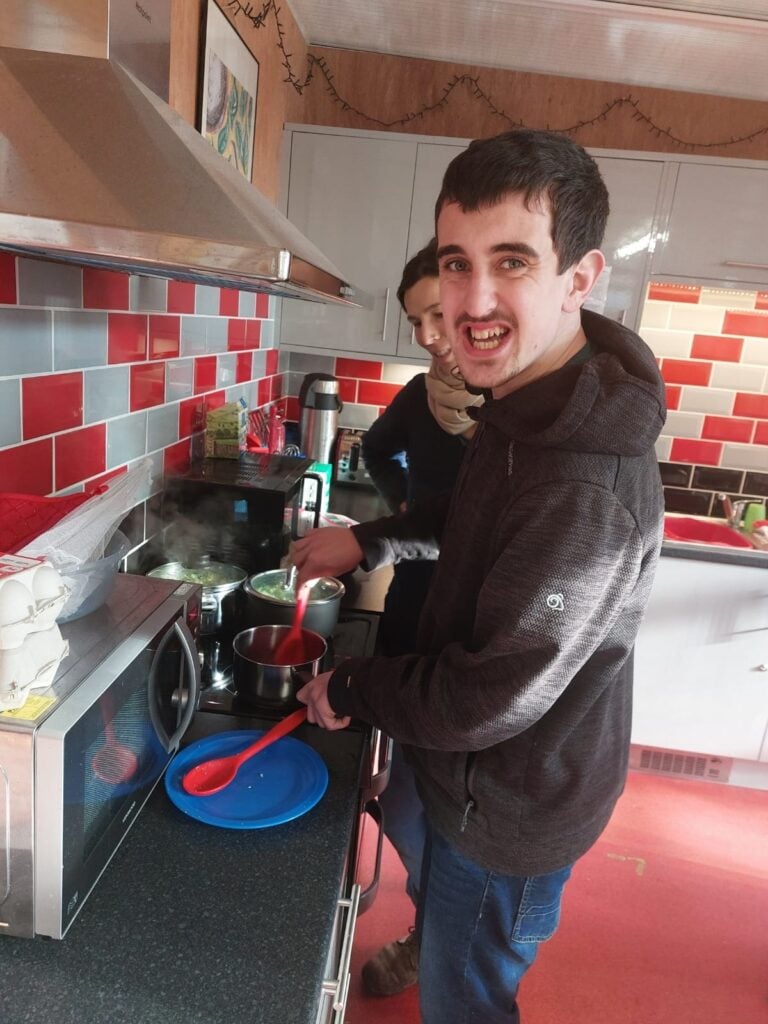 Young man in a home kitchen stirs a saucepan with support worker looking on
