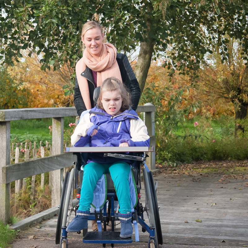 outdoor setting, young woman pushes a girl in a wheelchair both facing the front and smiling