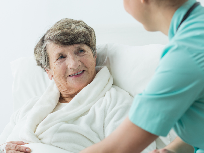 A nurse in light clothing sits beside an older woman in a hospital bed, offering support as the woman smiles warmly. Image to go with an article about the Dame Louise Casey review.